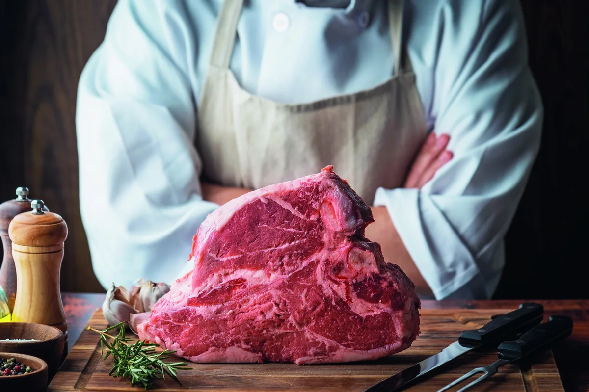 T-Bone-Steak_AdobeStock T-Bone Steak auf olzbrett, dahinter Metzger mit verschränkten Armen..