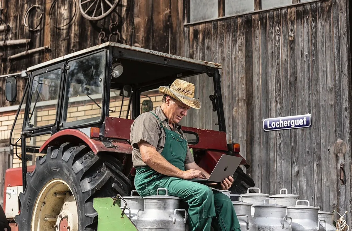 Landwirt sitzt auf Milchkannen und schreibt auf einem Notebook.